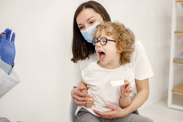 Little boy in eyeglasses is scared and screaming. Male doctor in white medical gown is going to do vaccination. Doctor and mother wearing face masks. - 写真・画像