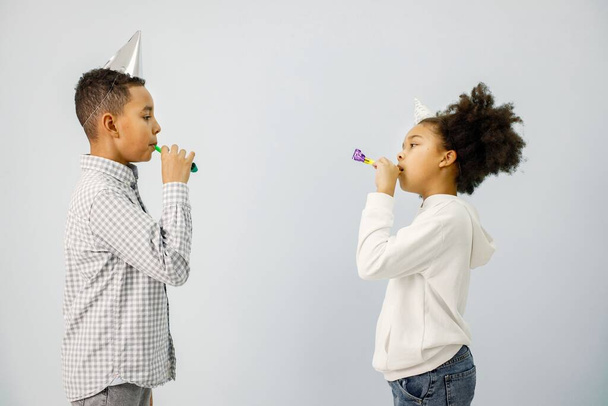 Boy and girl standing isolated on white background. Children blownig party horns. Black boy and girl wearing birthday cone hat. - Φωτογραφία, εικόνα