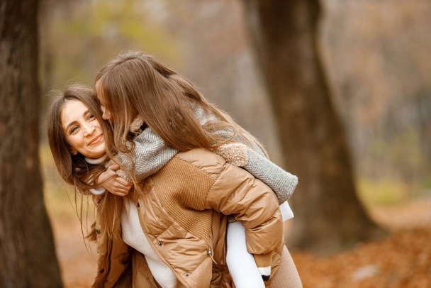 Young woman and little girl in autumn forest. Woman carry her daughter piggyback. Girl wearing fashion grey dress with a jacket. - Photo, Image