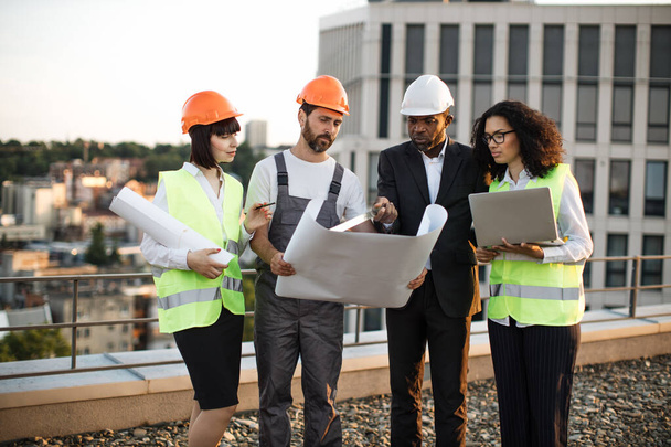 Group of four multiracial people in suits and helmets brainstorming for common project of new real estate building. Construction team working with blueprints and modern laptop in fresh air. - Photo, image