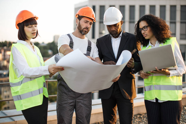 Diverse group of architects looking at construction diagram and discussing settlement of terraces on residential complex. African american woman holding portable laptop for entering data in program. - Foto, Imagen