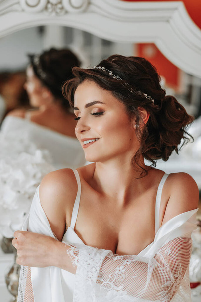 tall bride with bare shoulders, open bust, in a room near a mirror. Great wedding hairstyle. lace robe. Portrait. Delicate wedding makeup - Foto, Bild