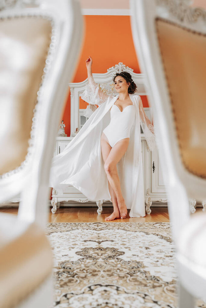 tall bride with bare shoulders, open bust, in a room near a mirror. Great wedding hairstyle. lace robe. Portrait. Delicate wedding makeup - Foto, Imagem