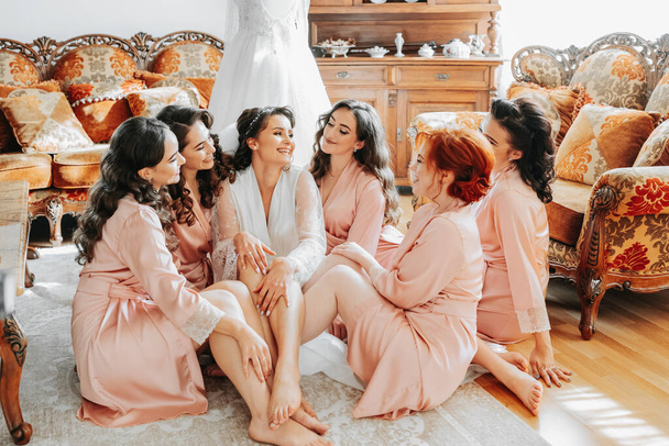 The bridesmaids look at the smiling bride. The bride and her fun friends are celebrating a bachelorette party on the floor near the wedding dress in matching dresses. Bride and friends in the room - Фото, изображение