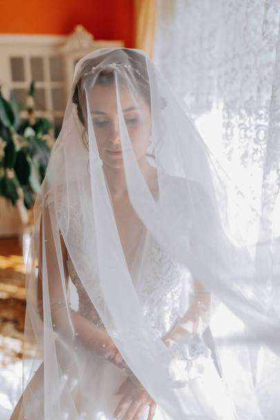 A brunette bride in a white dress holds her wedding bouquet, poses. French manicure. Open shoulders. Beautiful hands. Long veil. Morning of the bride. Details - Fotoğraf, Görsel