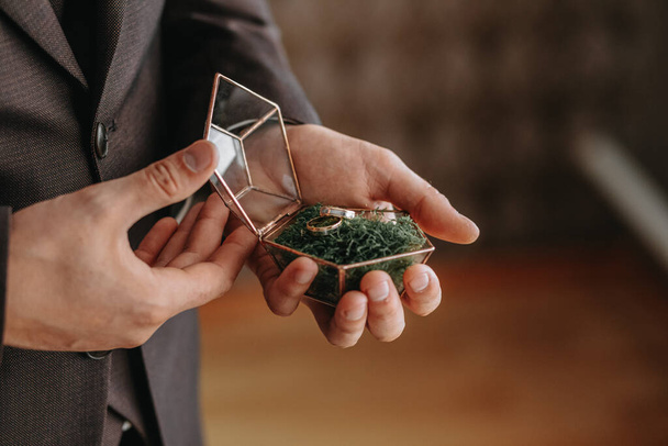 Wedding gold rings on a decorative glass box with wooden green moss, standing in the hands of the groom, close-up. Jewelry concept - Fotografie, Obrázek