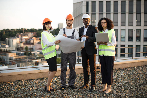 Full length portrait of multiethnic work team standing on terrace with amazing city view and looking at camera. Four international urban planners dressed in protective wear holding layouts and laptop. - Φωτογραφία, εικόνα