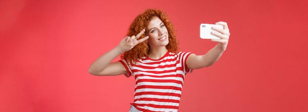 Стильная беззаботная красивая рыжая кудрявая женщина наслаждаются летними каникулами показывают победу жеста жеста глаза наклон головы милый запись видео провести смартфон с селфи красный фон. Копирование пространства - Фото, изображение