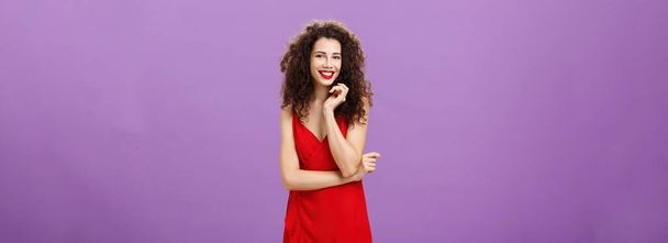 Gorgeous lady. in red feeling shy and awkward being invited for dance standing timid and feminine over purple background touching strand of curly hair smiling broadly and crossing arm over chest. - Foto, Imagem