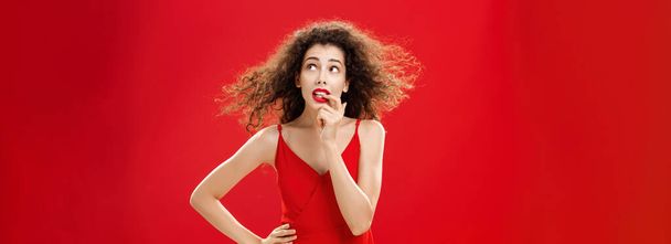 Waist-up shot of concerned and worried insecure cute female with curly hairstyle in red evening dress biting finger looking troubled or questioned at upper right corner with hand on waist. Copy space - Photo, Image