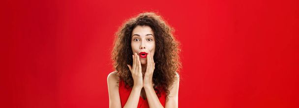 Charmed and intrigued curious good-looking rich woman in elegant dress with make-up and curly hairstyle folding lips in wow sign holding hands on face and smiling from excitement over red background - Foto, Imagem