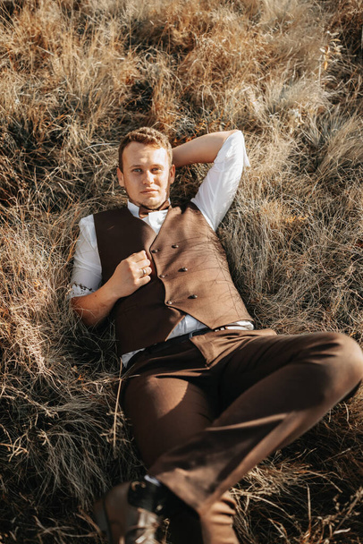 portrait of a stylish groom on a background of dry autumn grass. the concept of a rural wedding in the mountains, happy bohemian newlyweds. man relaxing lying on the grass - Fotó, kép