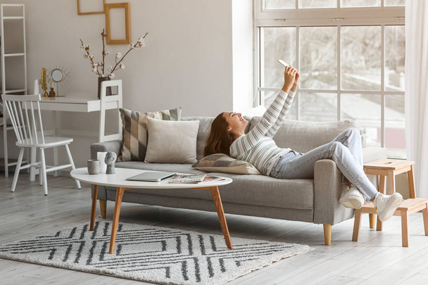 Young woman with mobile phone taking selfie on grey couch in living room - Foto, immagini