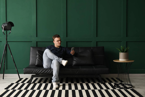 Jongeman met mobiele telefoon op zwarte bank in de woonkamer - Foto, afbeelding
