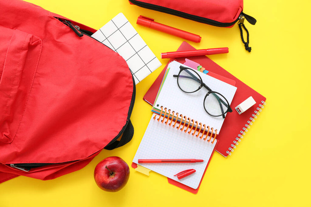 Mochila escolar roja con cuadernos, anteojos y estuche de lápiz sobre fondo amarillo - Foto, imagen