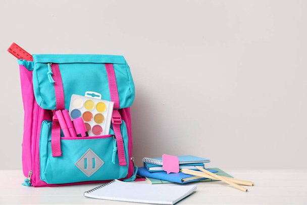 Кольоровий шкільний рюкзак з блокнотами, акварелями та маркерами на білому дерев'яному столі біля стіни - Фото, зображення