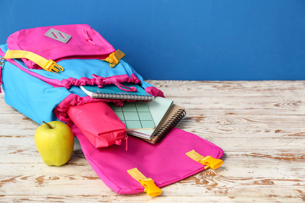 Kleurrijke schoolrugzak met notitieboekjes, etui en appel op houten tafel bij blauwe muur - Foto, afbeelding