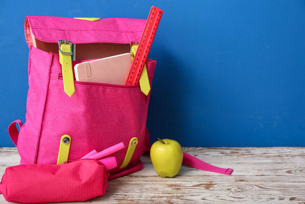 Рюкзак розовой школы с пенал, маркеры и яблоко на деревянном столе рядом с синей стеной - Фото, изображение