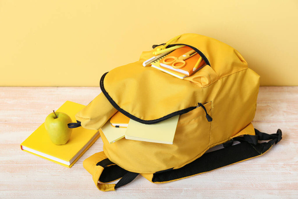 Sac à dos scolaire jaune avec cahiers, pomme et ciseaux sur table en bois près du mur - Photo, image
