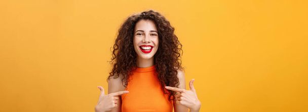 Kıvırcık saçlı, kırmızı rujlu, gururlu ve mutlu bir kadının turuncu arka plandaki başarısından bahsederken çekilmiş belden yukarı pozu. Boşluğu kopyala - Fotoğraf, Görsel