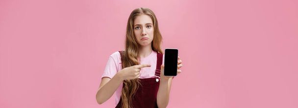 Lifestyle. Sad gloomy young female with cute wavy natural hairstyle showing smartphone screen pointing at gadget with index finger making upset face frowning displeased feeling regret after buying - Foto, Imagen
