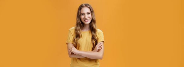 Životní styl. Portrét laskavé a přátelské půvabné mladé studentky v ležérním tričku s vlnitými přírodními dlouhými vlasy držící ruce zkřížené na prsou s širokým a bezstarostným úsměvem na kameru nad oranžovou - Fotografie, Obrázek