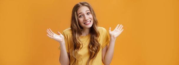 Mädchen halten sich achselzuckend mit erhobenen und zur Seite gespreizten Händen aus dem Geschäft heraus, dummes, trauriges Lächeln posiert über orangefarbenem Hintergrund, ahnungslos und verwirrt in lässigem T-Shirt, das keine Ahnung vom Thema hat. - Foto, Bild