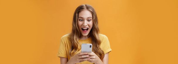Lifestyle. Excited and thrilled charismatic young happy girl in yellow t-shirt smiling with opened from joy mouth holding smartphone staring at cellphone screen astonished carried away with cool phone - Photo, Image