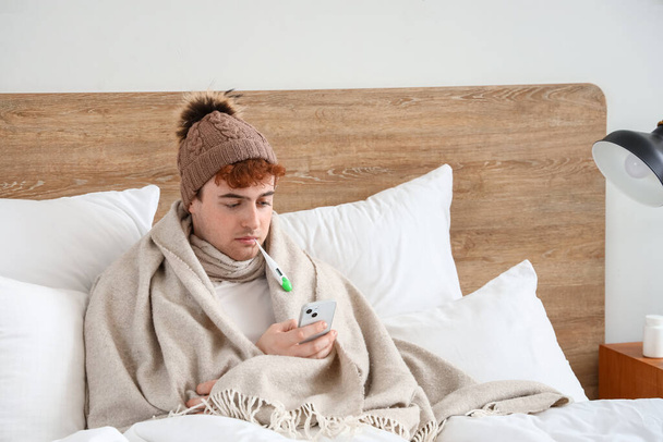 Jeune homme malade avec thermomètre utilisant le téléphone portable dans la chambre - Photo, image