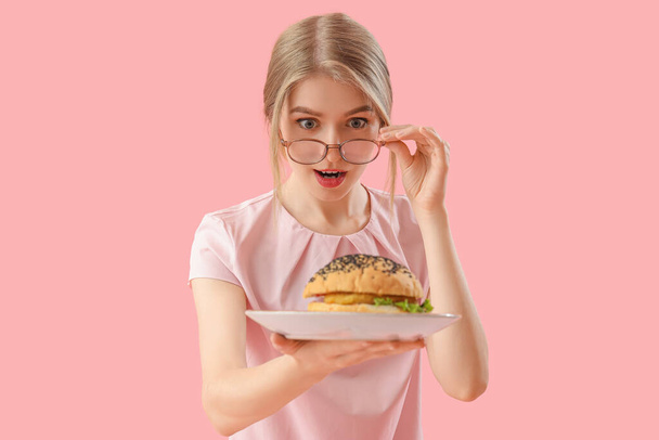 Shocked young woman with tasty burger on pink background - Фото, зображення