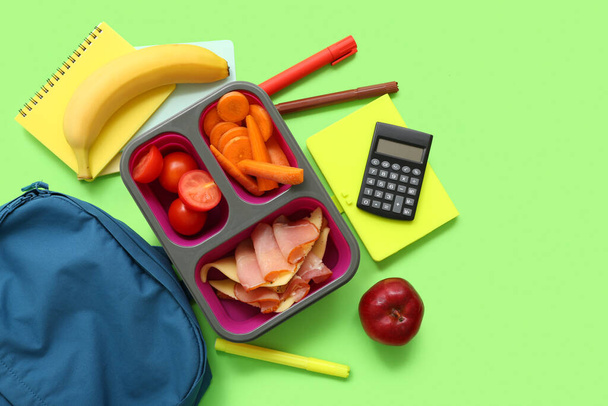 Plecak, papeteria i pudełko na lunch ze smacznym jedzeniem na zielonym tle - Zdjęcie, obraz