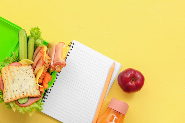 Stationery, drink and lunch box with tasty food on yellow background - Photo, Image