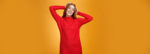 Lifestyle. Romantic and sensual cute ginger girlfriend in elegant warm red winter dress holding hands behind head relaxed and carefree smiling delighted tilting head enjoying leisure and holidays. - Foto, afbeelding