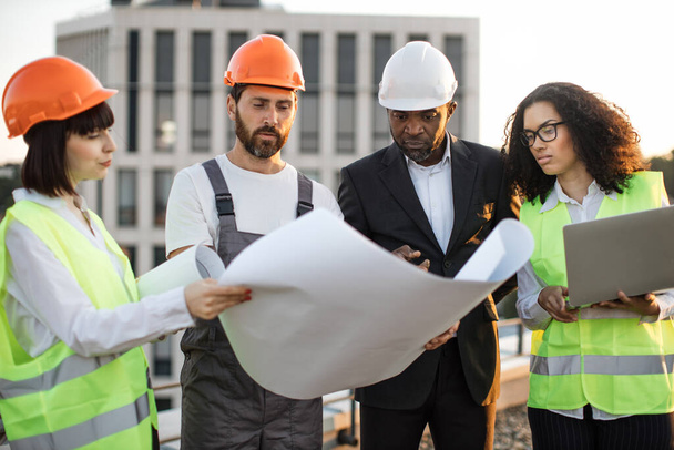 Diverse group of architects looking at construction diagram and discussing settlement of terraces on residential complex. African american woman holding portable laptop for entering data in program. - Photo, image