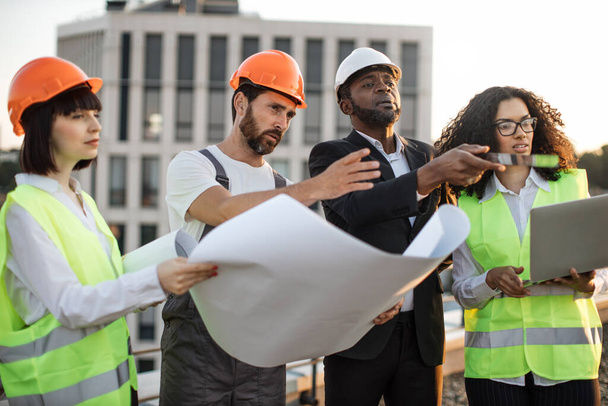 Diverse group of architects looking at construction diagram and discussing settlement of terraces on residential complex. African american woman holding portable laptop for entering data in program. - Foto, immagini