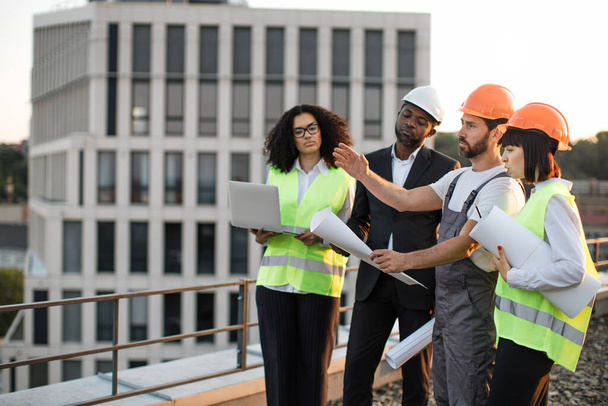 Four multiracial engineers with technical drawings and laptop discussing place for construction of new multi-apartment complex. Workman talking and pointing with hand while others listening carefully. - Photo, image