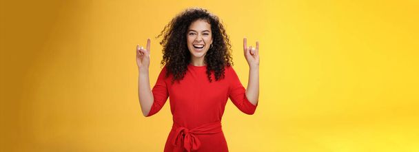 Rock n roll live. Charismatic excited young curly-haired woman in red dress feeling awesome as waiting concert of favorite band showing rock gestures with raised hands and smiling thrilled and happy. - Photo, Image