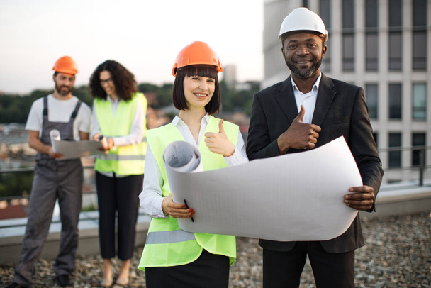 Attractive caucasian lady and african american man holding construction plan together and happily smiling at camera, showing thumbs up. - Photo, image
