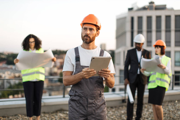 Diverse team of international construction inspectors in vests and helmets attending building area and looking at blueprints. Focused engineer with digital device in hands monitoring working process. - Foto, Imagen