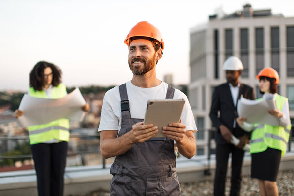 Diverse team of international construction inspectors in vests and helmets attending building area and looking at blueprints. Focused engineer with digital device in hands monitoring working process. - Foto, Imagen