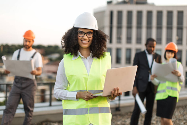 Portrait of african american female urban planner in protective wear and glasses holding remote computer and looking at camera. Diverse architects reviewing building blueprints on roof terrace. - Φωτογραφία, εικόνα