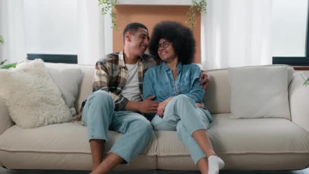 Счастливая семья афро-американская пара влюбленная обнимается на диване дома, проводит время вместе любящий мужчина, любящий мужчину, муж, парень, жена, подружка обнимает обнимающих домовладельцев, арендаторов недвижимости - Кадры, видео