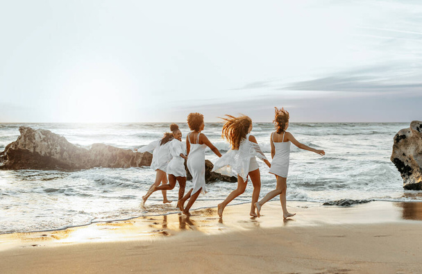 Happy young female friends having fun and celebrating hen party while running on the beach at the sunset, enjoying evening at coastline, free space - 写真・画像