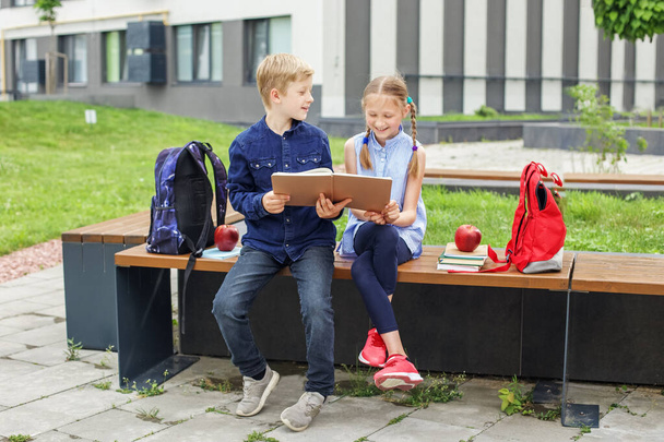 Возвращайся в школу. Двое детей, мальчик и девочка, читают книги вместе и разговаривают, сидя на скамейке в школьном дворе. - Фото, изображение
