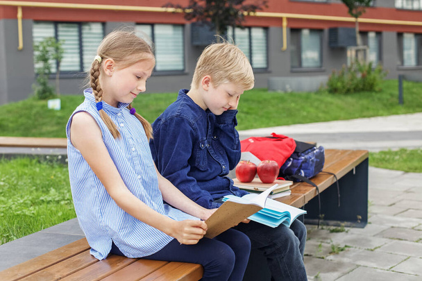 Τα παιδιά διαβάζουν βιβλία που κάθονται στον πάγκο στην αυλή του σχολείου. Πίσω στο σχολείο. Έναρξη των σχολικών μαθημάτων. - Φωτογραφία, εικόνα