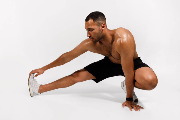 Fitness training. Atletische Afro-Amerikaanse man doet Hamstring Stretches Oefening Over White Studio Achtergrond. Sport Guy met Shirtless Torso stretching Leg Muscles. Totale lengte - Foto, afbeelding