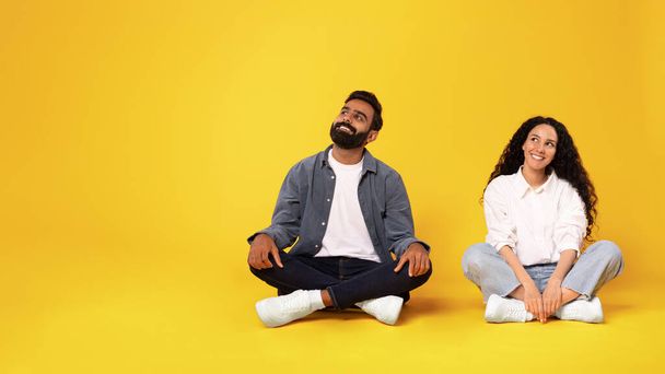 Great Offer. Smiling arabic couple in casual sitting and looking aside at space for advertisement, posing together on yellow studio background. Banner with positive spouses advertising your text - Photo, Image