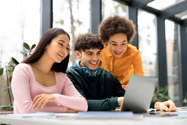 Szczęśliwi międzynarodowi studenci studiujący w przestrzeni coworkingowej, cieszący się wynikami swojego projektu, patrzący na ekran laptopa i uśmiechający się, siedzący razem w pomieszczeniach - Zdjęcie, obraz