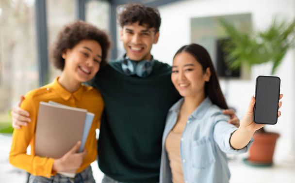 Tři šťastní různí studenti přátelé objímající a asijské dáma ukazující prázdný displej mobilu, reklama vzdělávací app, stojící na chodbě vysoké školy, mockup - Fotografie, Obrázek