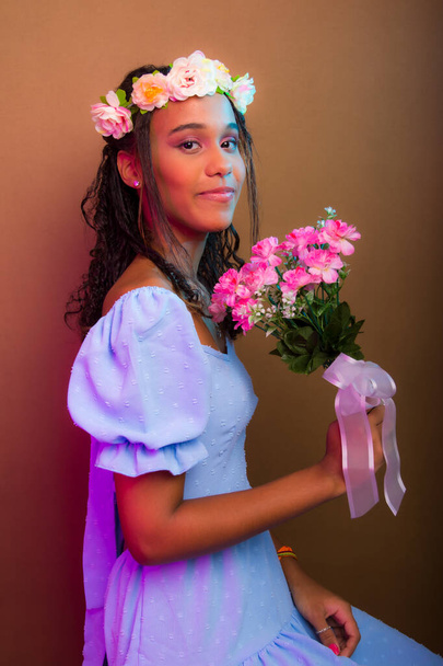 Portrait of beautiful girl, teenager, wearing blue, with tiara on her head and holding bouquet of flowers. Isolated on brown background. - Foto, Imagem